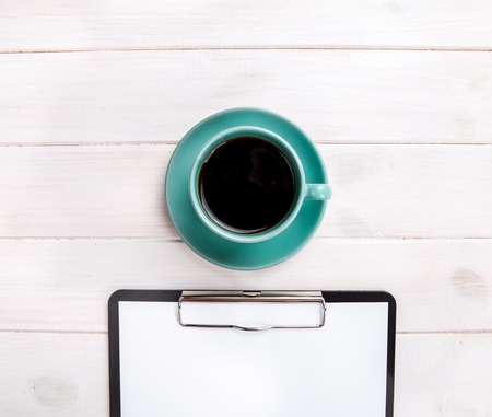 Clipboard with blank page and cup of coffee on wooden desk. View from above.
