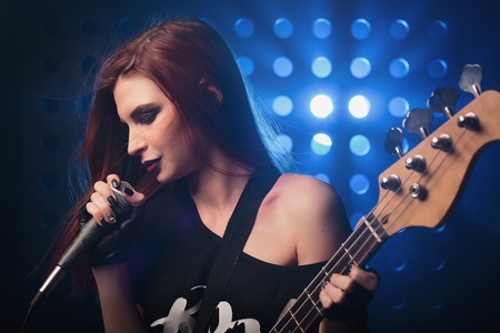 Beautiful girl with a microphone and a guitar in hand on stage singing a song in rock style, closeupの写真素材