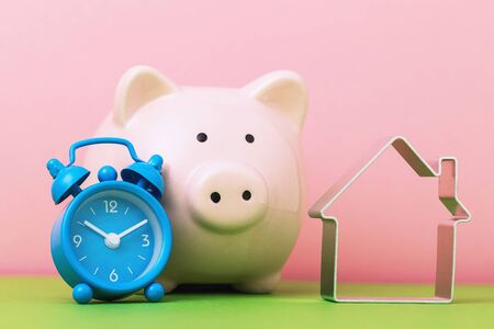 Its time to save up for real estate. Alarm clock, piggy bank and object in the shape of a house. Concept of accumulating money for housingの写真素材