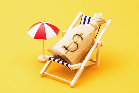 Wooden bag with a dollar symbol in a deck chair and a parasol on a yellow background. Concept on the cost of vacationの写真素材