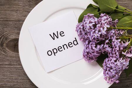 Note with text in a white plate on a wooden table and a branch of lilac. The concept of opening a restaurant business in connection with restrictive measures due to the Covid-19 pandemicの写真素材
