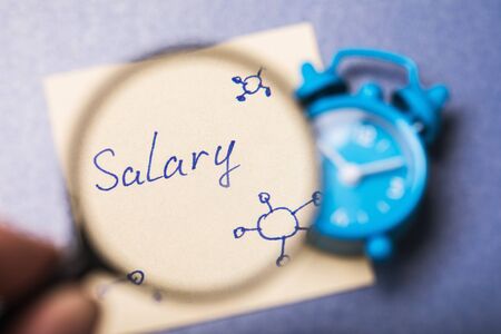 Hand with a magnifier and an alarm clock with a note on the table. Concept on the effect of coronavirus on peoples salariesの写真素材