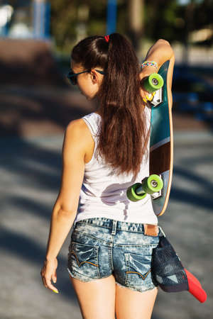 Girl with a skateboard in hands goes to the sports field on a summer sunny eveningの写真素材