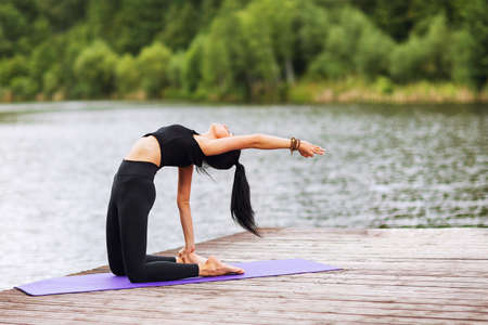 Brunette girl with long hair in a black sports suit does exercises, sitting on a mat laid out on wooden boards by the riverの写真素材