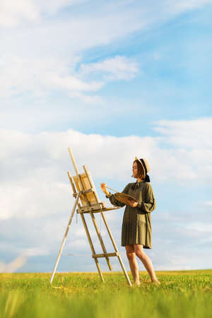 Young woman artist paints a picture outdoors on a warm summer evening. Model posing in dress barefootの写真素材