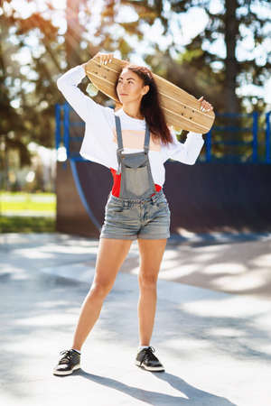 Portrait of a beautiful young woman with a longboard in her hands in the park on a sunny summer...