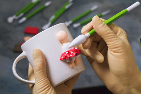 A master makes a figurine of a person on a ceramic mug made of polymer clay, close-upの写真素材