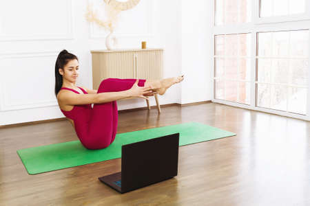 Young woman in sportswear practicing yoga, exercising at home with an online trainer, doing boat pose, Paripurna Navasana. Concept on the topic of online learning.の写真素材