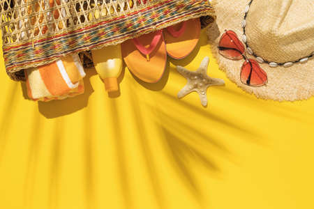 Straw bag with beach holiday accessories on yellow background and palm leaf shadow with copy space,...