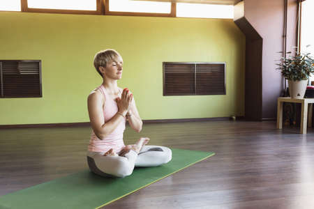 Attractive blonde woman in sportswear, practicing yoga, is engaged in meditation in the lotus position with namaste, on the rug in the studio.の写真素材