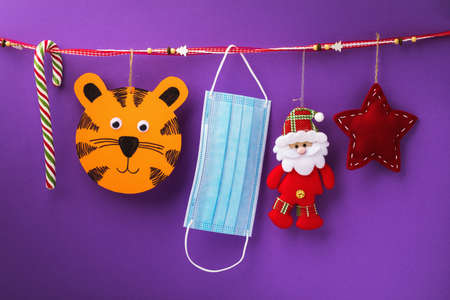 Symbol of 2022, Christmas tree toys and a medical mask hang on a decorative ribbon on a colored...