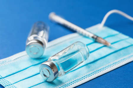 Two vials with a solution for injection, a disposable syringe and a medical mask on a blue...