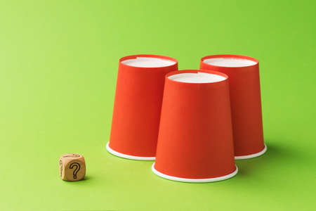 Three orange paper cups and one wooden cube with a question mark symbol, the concept of gambling and its harmの写真素材