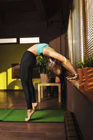A woman performs the exercise Urdhva Dhanurasana, bridge pose from a standing position, resting her hands on the windowsill, stands on the rug in the studioの写真素材