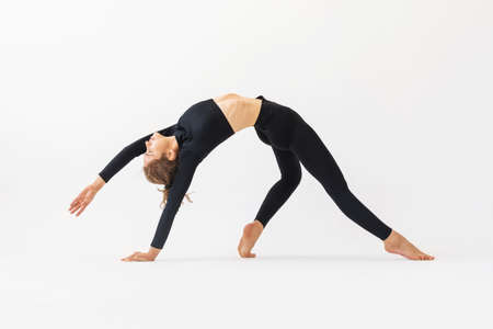 A young woman practicing yoga performs the exercise of Kamatkarasana, the pose of a dancing dog, trains in black sportswear on a white backgroundの写真素材