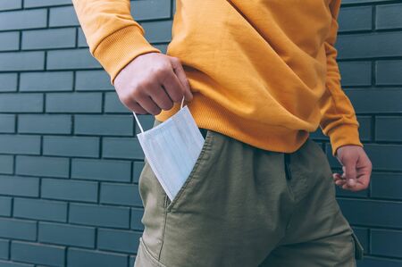 A guy in a yellow sweater is walking against a brick wall and takes a medical mask from his pocket....