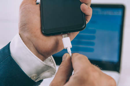 Man charges a smartphone through a laptop on a white tableの写真素材