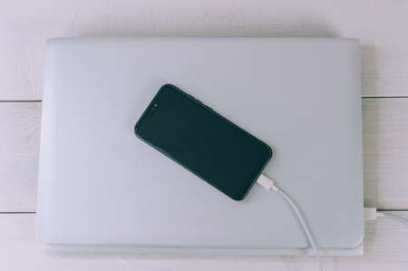 The smartphone is charging from a laptop, on a white, wooden tableの写真素材