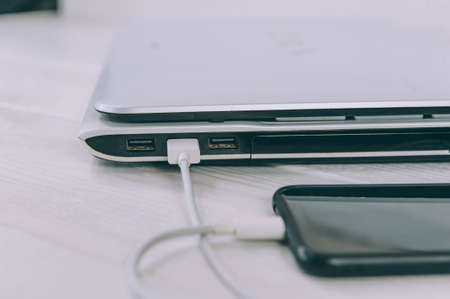 The smartphone is charging from a laptop, on a white, wooden tableの写真素材