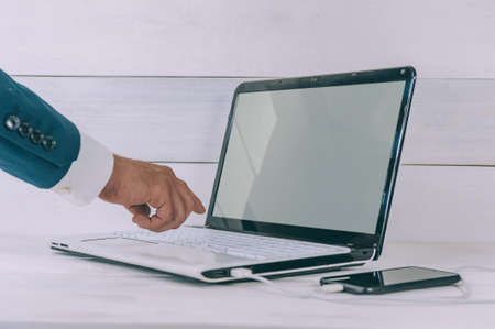 Businessman works on a laptop and charges a smartphone on a white, wooden tableの写真素材