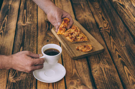 The guy holds pizza on a wooden table against the background of a cup of coffee. Fast food cooked at...