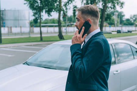 A businessman in a blue jacket is talking on the phone on the background of the carの写真素材