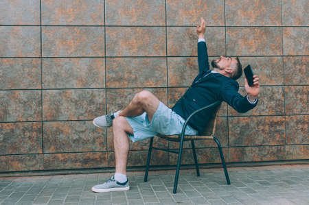 Businessman in a blue jacket and shorts, sitting on a chair, with a smartphone in his handの写真素材
