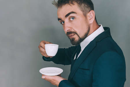 Businessman in a jacket drinks and holds in his hand a cup with a coffee on a gray backgroundの写真素材
