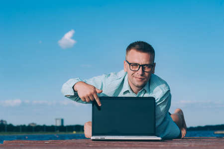 A man in glasses and a shirt working on a laptop in the fresh air, against the background of a riverの写真素材