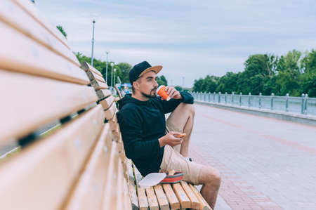 Hipster guy was sitting on a bench and having dinner with toast with coffee in a glass. Fast food in the fresh airの写真素材