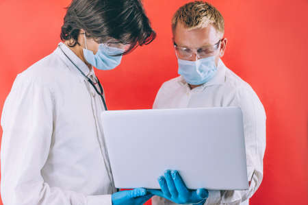 Two doctors with blue gloves in masks on the face with a laptop on a red backgroundの写真素材