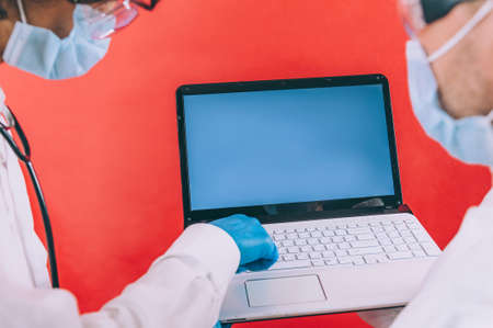 Two doctors with blue gloves in masks on the face with a laptop on a red backgroundの写真素材