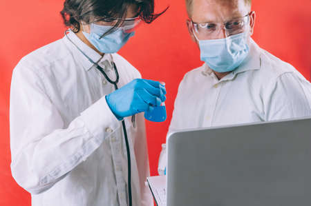 Two doctors with blue gloves in masks on the face with a laptop on a red background