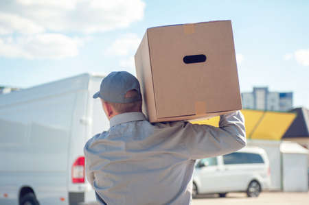 A man in a gray suit in his hands with a cardboard box against the background of the van. Concept on the topic of postal deliveryの写真素材