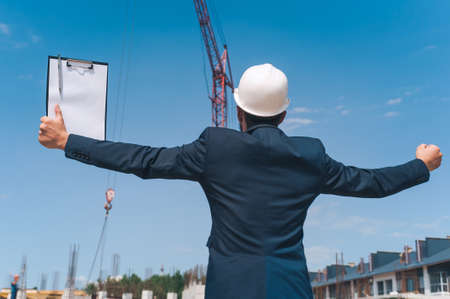 The chief in a jacket and a construction helmet with documents in his hands, against the background of a tower crane and the sky with clouds. House construction inspectionの写真素材