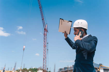 The chief in a jacket and a construction helmet with documents in his hands, against the background of a tower crane and the sky with clouds, is talking on a smartphoneの写真素材
