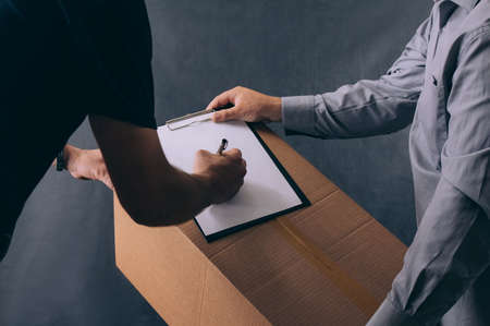 A man with a cardboard box and documents in his hands, hands over the parcel to the client. Delivery service concept on dark backgroundの写真素材