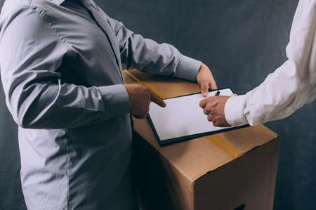 Man with a cardboard box gives a package to a client. Delivery service concept on dark background