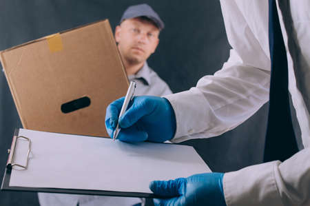A man with a cardboard box and documents in his hands, hands over the parcel to the client. Delivery service concept on dark backgroundの写真素材