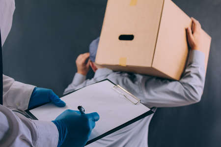 A man with a cardboard box and documents in his hands, hands over the parcel to the client. Delivery service concept on dark backgroundの写真素材