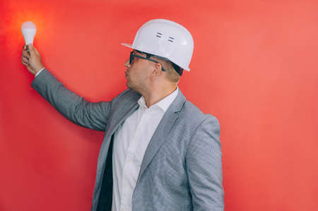 The chief in a construction helmet holding a light bulb in his hand on a red backgroundの写真素材