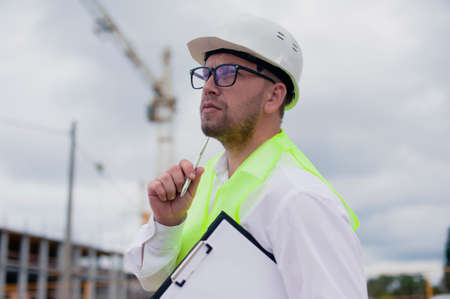 Chief in a vest and a construction helmet with documents in his hands, against the background of a...