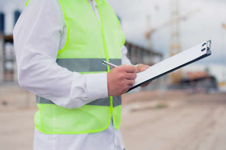 Chief in a vest and a construction helmet with documents in his hands, against the background of a...
