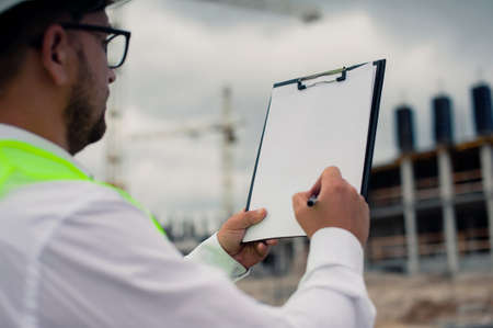 Chief in a vest and a construction helmet with documents in his hands, against the background of a tower crane and the sky with clouds. House construction inspectionの写真素材