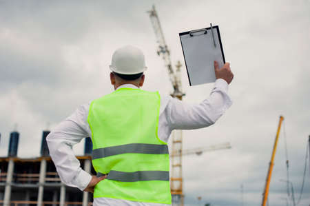 Chief in a vest and a construction helmet with documents in his hands, against the background of a tower crane and the sky with clouds. House construction inspectionの写真素材