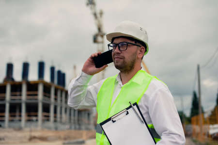 The chief in a jacket and a construction helmet with smartphone in his hands, against the background of a tower crane and the sky with cloudsの写真素材
