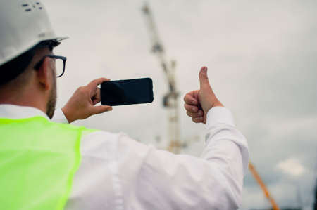 The chief in a jacket and a construction helmet with smartphone in his hands, against the background of a tower crane and the sky with cloudsの写真素材