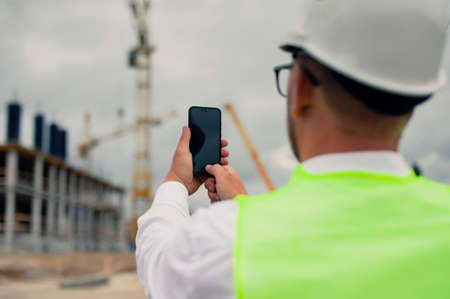 The chief in a jacket and a construction helmet with smartphone in his hands, against the background of a tower crane and the sky with cloudsの写真素材