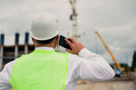 The chief in a jacket and a construction helmet with smartphone in his hands, against the background of a tower crane and the sky with cloudsの写真素材