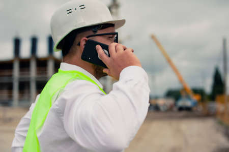 The chief in a jacket and a construction helmet with smartphone in his hands, against the background of a tower crane and the sky with cloudsの写真素材